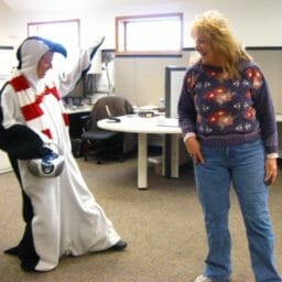 A laughing blonde woman in an office, next to a woman dressed in a penguin costume.