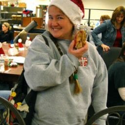 A smiling woman in a Santa hat and gray sweatshirt at an office, holding a Christmas gift jar.