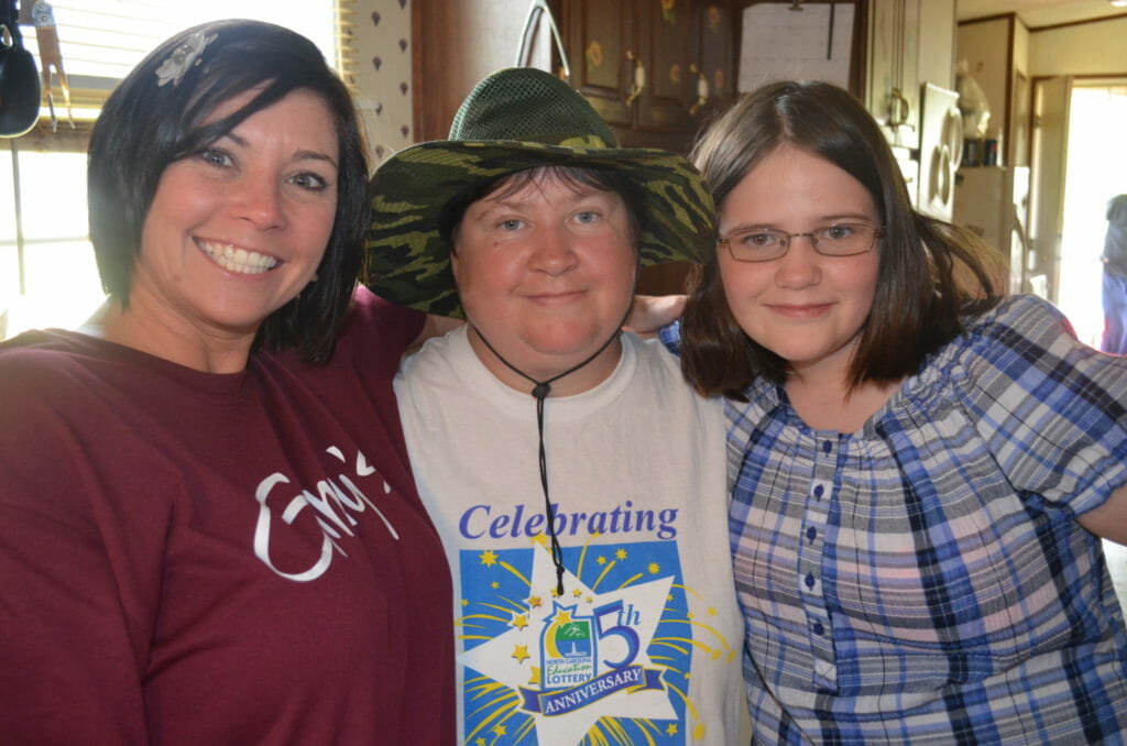 Three smiling women in a home kitchen, one in a Ginny's T-shirt.