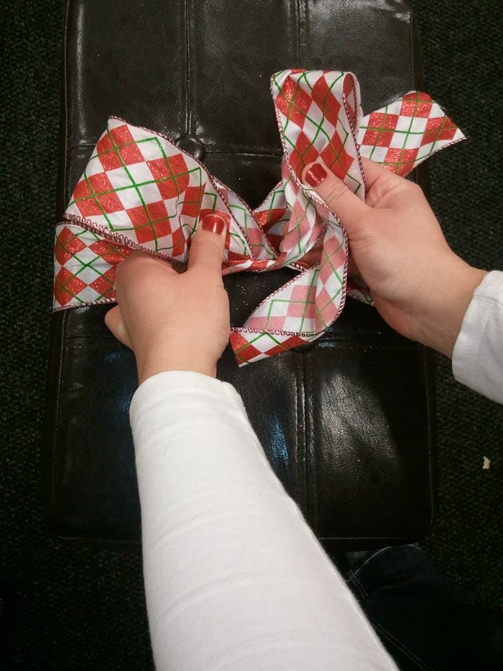 A woman's hands tying a red, green, and white plaid ribbon bow around a brown leather stool.
