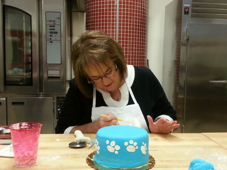 Ginny, a brunette woman, placing white fondant paw print cutouts on the side of a blue layer cake.