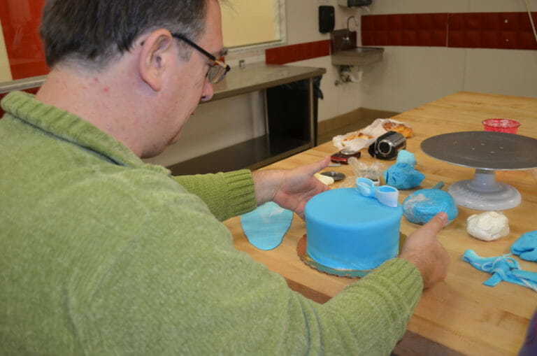 A man in glasses at a bakery table, decorating a layer cake with blue fondant icing.