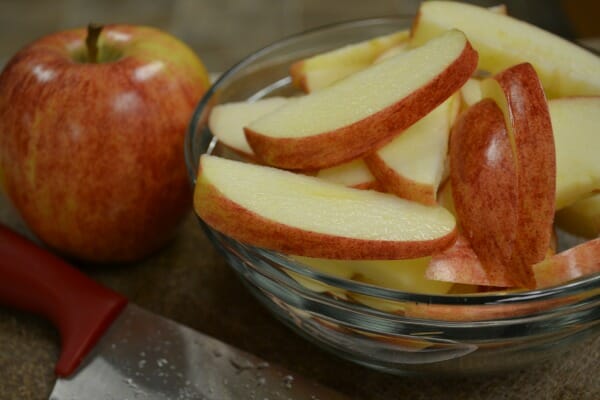 A red apple next to a clear bowl of slender apple slices and a red-handled knife.