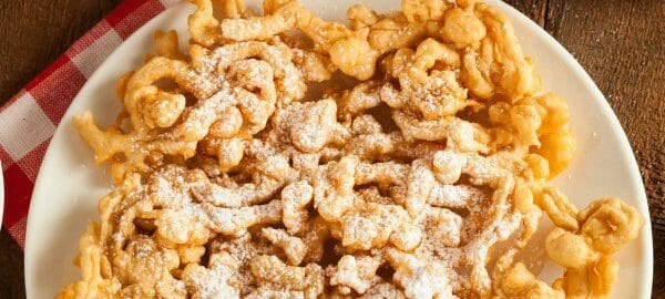 A white plate filled with Funnel Cakes dusted with powdered sugar, with a red check napkin on a picnic table.