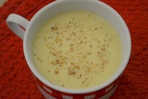A red and white check mug on a red napkin, filled with Eggless Eggnog sprinkled with nutmeg.