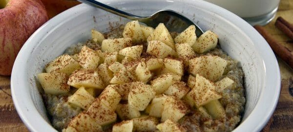 A white bowl filled with Cinnamon Steel-Cut Oatmeal with apple chunks, next to a glass of milk and two red apples.