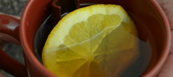 Close-up of a red mug filled with Mulled Apple Cider, with a lemon wedge and a cinnamon stick.