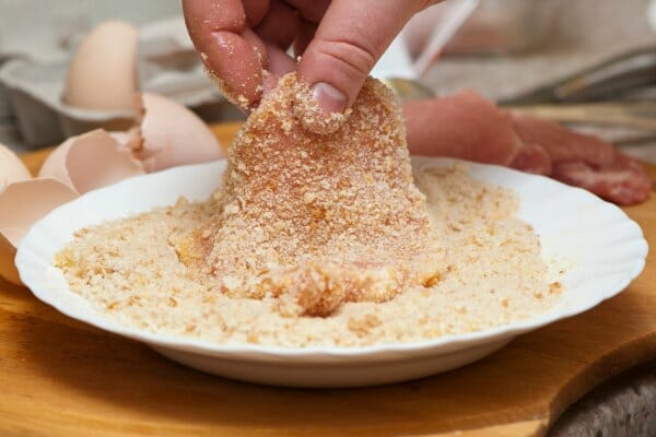 A hand dredging a pork tenderloin in a plate filled with breading crumbs.