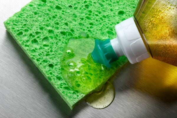 A bottle filled with gold dish detergent being squeezed out onto a green sponge.