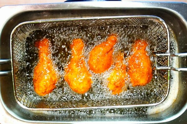 Five chicken legs sizzling in a deep fryer filled with bubbling oil.