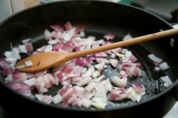 A hot black cast iron frying pan filled with chopped red onions, being stirred by a wooden spoon.