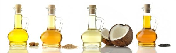 Four different shades of golden oil in glass cruets, with nuts, seeds, and a coconut, depicting the oil sources.