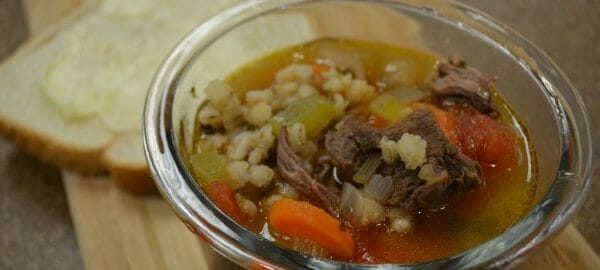 An individual serving of beef barley soup in a clear glass bowl, with a slice of buttered bread.
