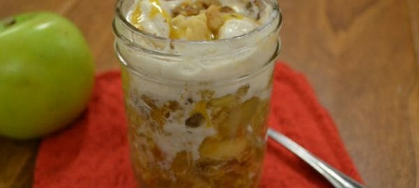 A serving of Apple Crisp in a clear jelly jar, with a spoon and a green Granny Smith apple nearby.