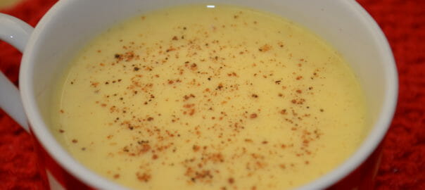 A red and white check mug on a red napkin, filled with Eggless Eggnog sprinkled with nutmeg.