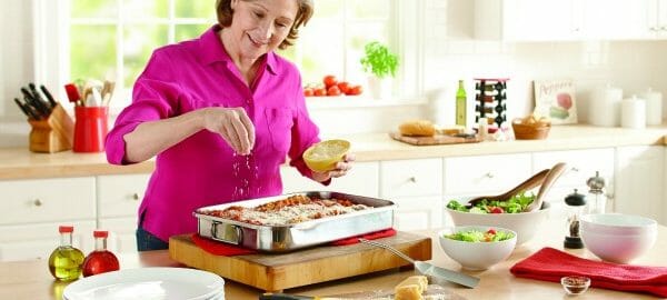 A woman in a fuschia blouse in a kitchen, sprinkling shredded cheese on a pan of lasagna, with a salad nearby.
