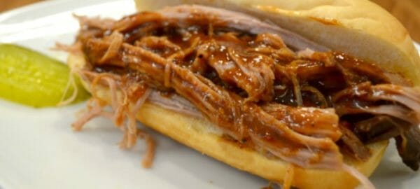 A white plate with barbeque pulled pork on a hard bun, with a dill pickle slice on the side.