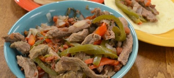 A blue bowl filled with Slow Cooker Fajitas next to a stack of soft taco shells, and a plate with a single serving.