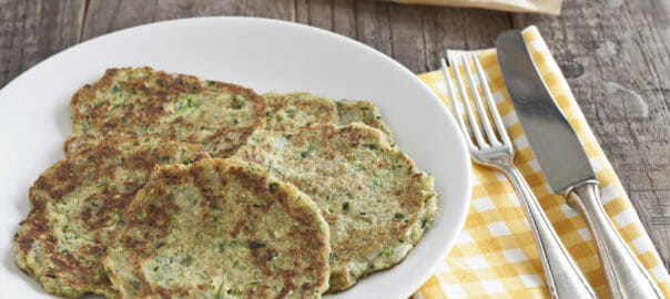 A white plate filled with zucchini patties, next to a fork and knife on a yellow check napkin.