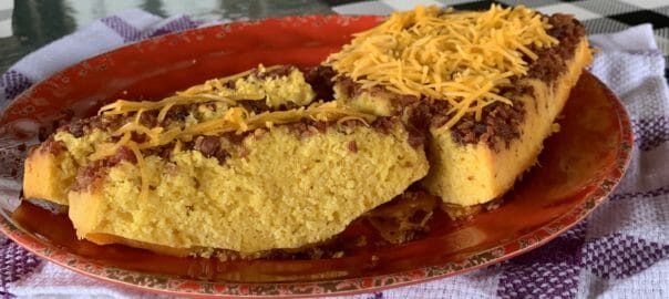Cornbread topped with shredded cheese and bacon, served on a decorative plate with a checkered tablecloth background.
