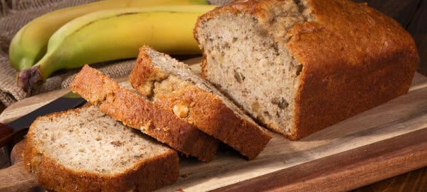 A loaf and three cut slices of Banana Bread on a wooden cutting board, next to two bananas.