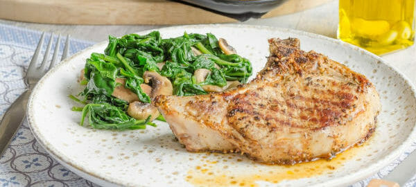 A white plate with a serving of Cider Pork Chops and mushroom spinach, with a glass of cider nearby.