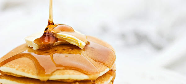 Maple syrup being poured on a stack of fluffy pancakes topped with butter pats.