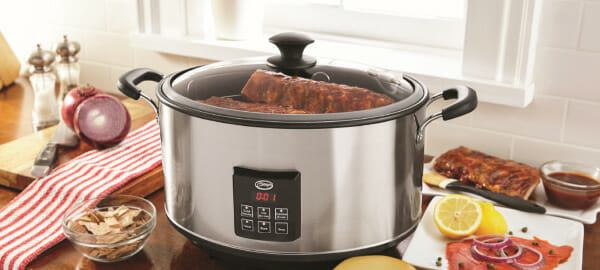A Ginny's brand slow cooker filled with smoked ribs, on a counter with cheese on a cutting board, and BBQ sauce.