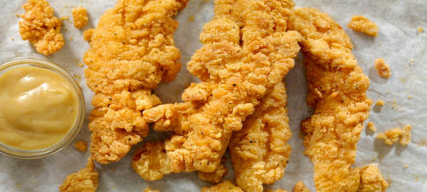 Four Crispy Chicken strips on parchment paper with a ramekin of Dijon mustard for dipping.