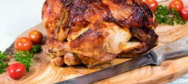 A whole browned Teriyaki Rotisserie Chicken on a wooden cutting board, with cherry tomatoes, parsley, and a knife.