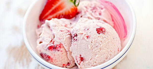 A white bowl filled with homemade strawberry ice cream, with a pink spoon and a fresh strawberry garnish.