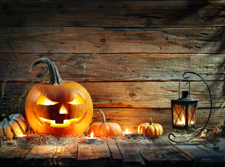 A lit Jack-o-Lantern on a rustic board porch, with small pumpkins, grasses, a lit lantern and tea lights.