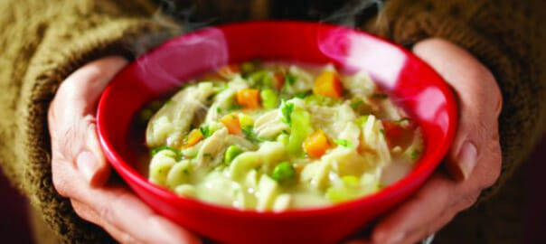 A person in a brown sweater holding a steaming red bowl filled with Creamy Chicken Noodle Soup.