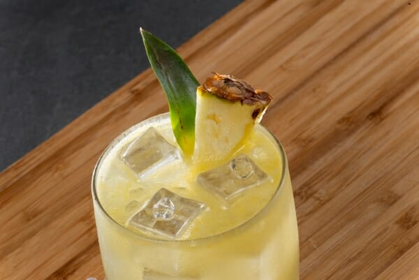 Refreshing yellow cocktail with ice, garnished with a slice of grilled pineapple and a green leaf, on a wooden surface.