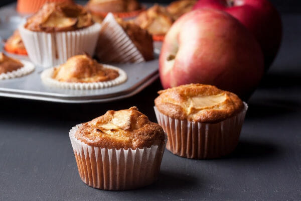 Freshly baked apple muffins with a golden top, surrounded by whole red apples and a muffin tray in the background.
