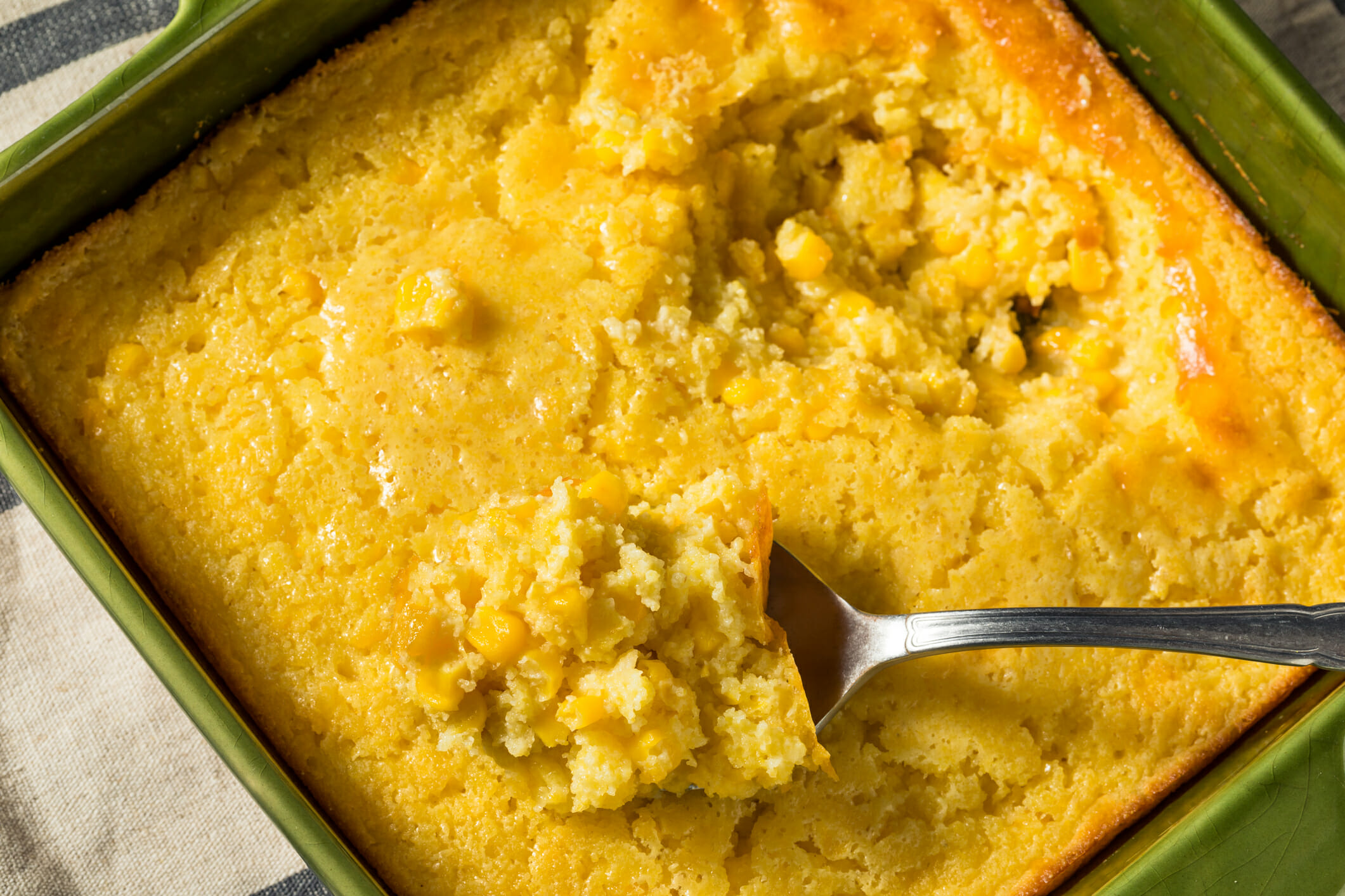 Golden cornbread with corn kernels, served in a green dish, with a spoonful taken out, resting on a striped cloth.