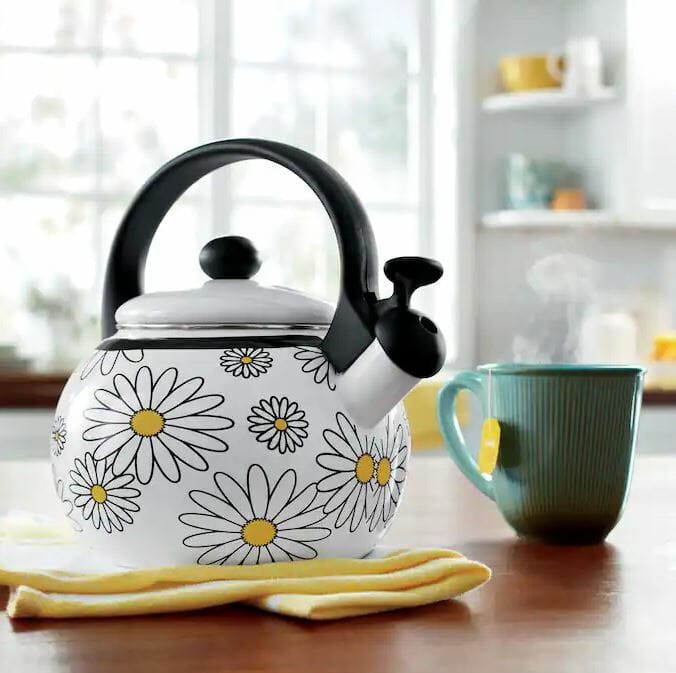 Whimsical daisy-patterned kettle beside a steaming cup on a wooden table, with a sunny kitchen backdrop.