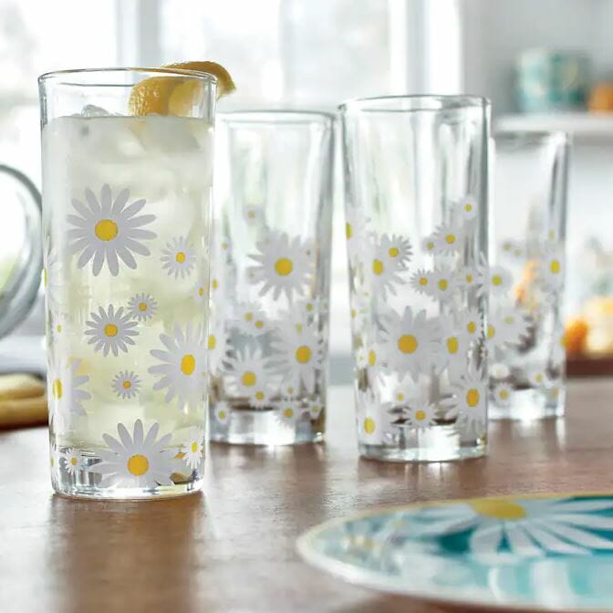 Tall glasses decorated with daisies, one filled with a lemon drink, on a wooden table with a colorful plate nearby.