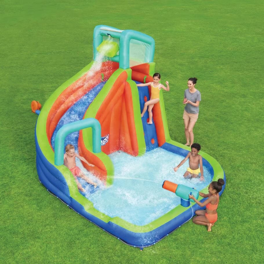 Colorful inflatable water slide with kids playing, splashing, and a woman supervising on a grassy lawn.