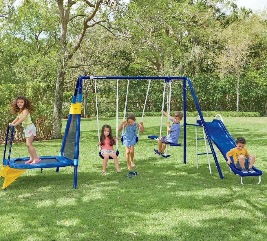 Five children play on a colorful swing set and slide in a sunny, green backyard.