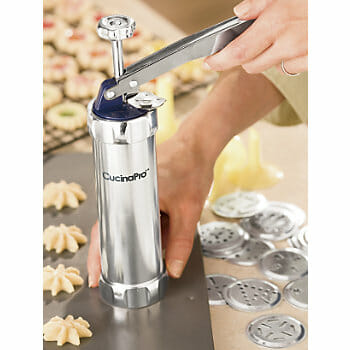 A silver cookie press with many design discs, with pressed cookies on a rack and a cookie sheet.