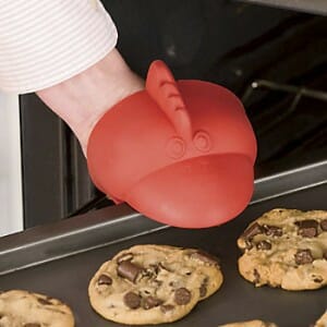 A hand in a red rooster-shaped silicone pot holder, taking chocolate chip cookies out of an oven.