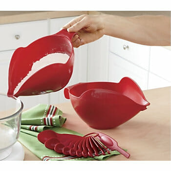 A hand pouring flour into a bowl from a large red measuring cup with spout, by a set of red measuring spoons. 