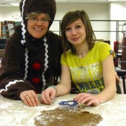Two smiling women cutting out gingerbread cookies on a floured counter, one in a cookie costume.