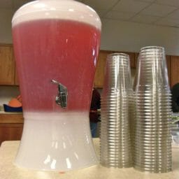 A clear drink dispenser filled with pink lemonade, with two stacks of clear plastic cups.