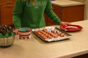 A foil-lined pan of bacon-wrapped sausages and a bowl of chocolate cake pops on a kitchen counter.