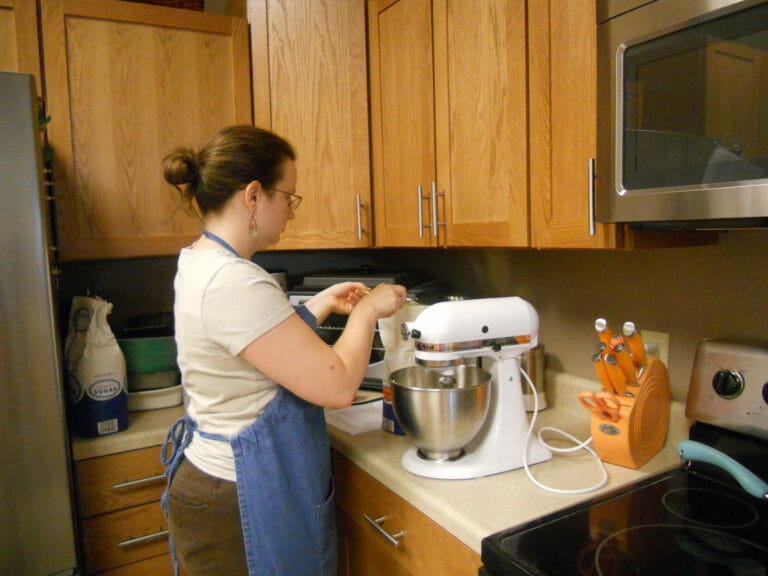 A woman wearing a denim apron in an office kitchen, mixing ingredients in a Kitchenaid mixer.