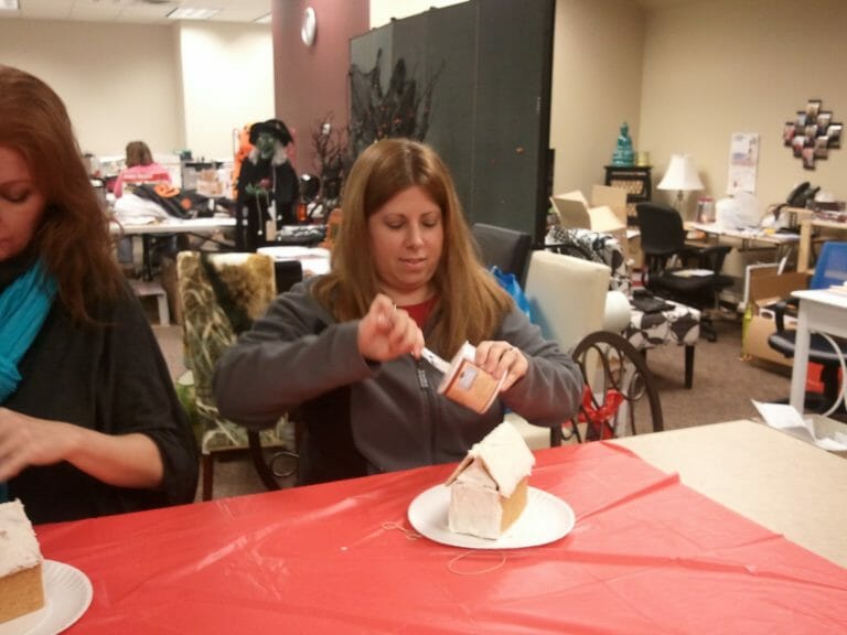 A woman icing a gingerbread house on a table in an office setting.