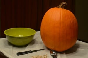 A pumpkin on a white plastic bag, with a green bowl, a knife, a scoop, and toothpicks.