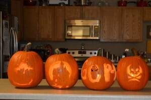 Four lit Jack-o-Lanterns on a kitchen counter, including a haunted house and a skull and crossbones design.